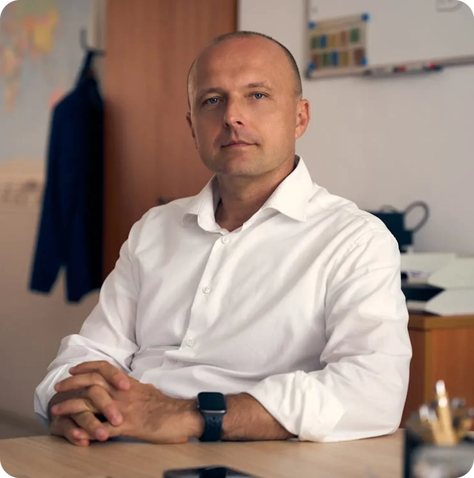 Serious middle-aged man in a white shirt sitting at a desk with hands clasped in an office setting.